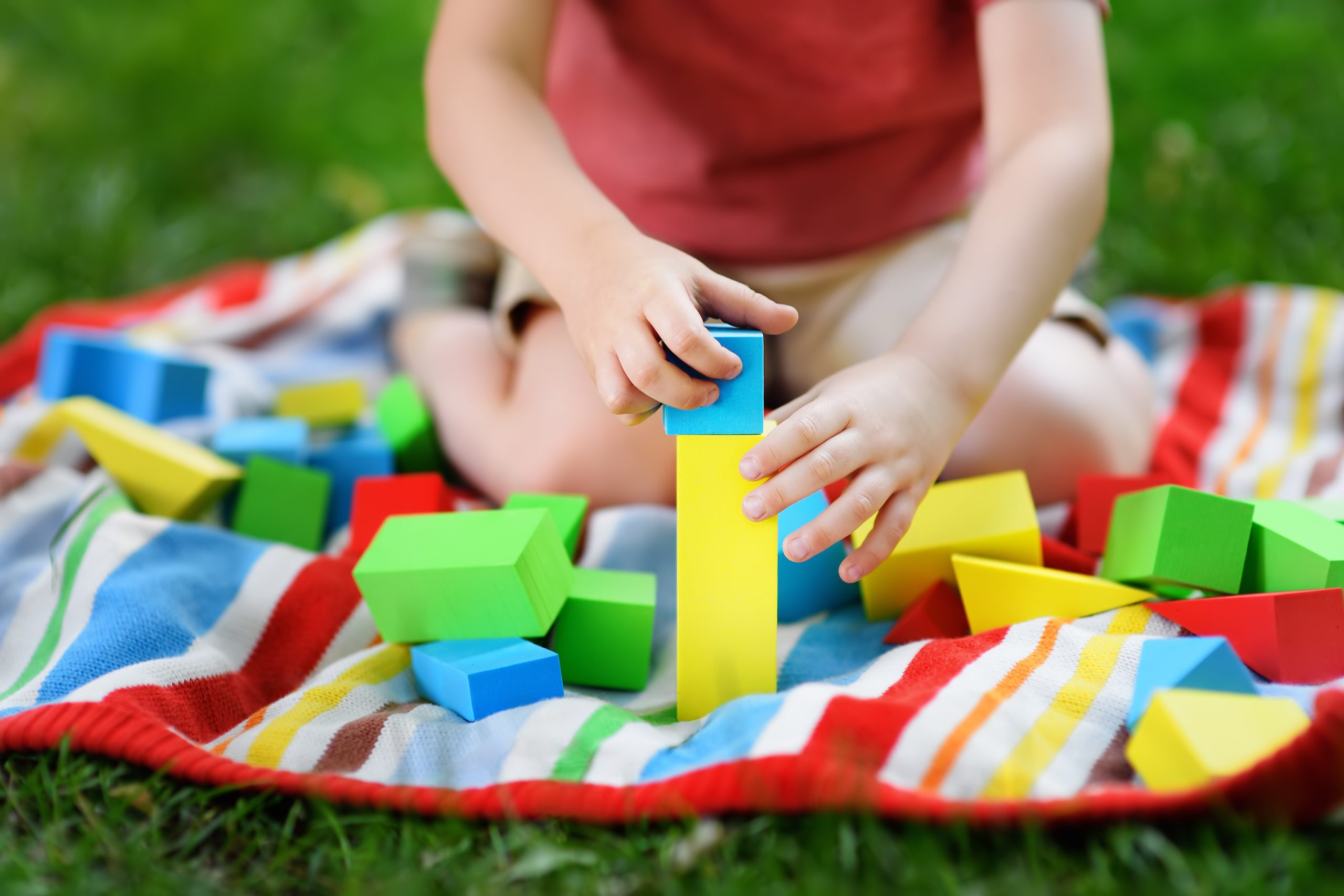 Cute preschooler boy playing with colorful cubes on the grass in summer park. Outdoors developing activity for kids. Family time with children on nature.