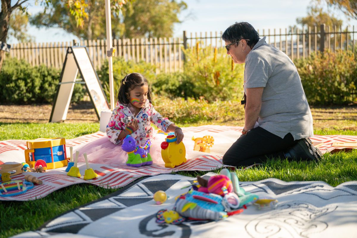 Toddler sitting on mat on grass, adult kneeling next to her.